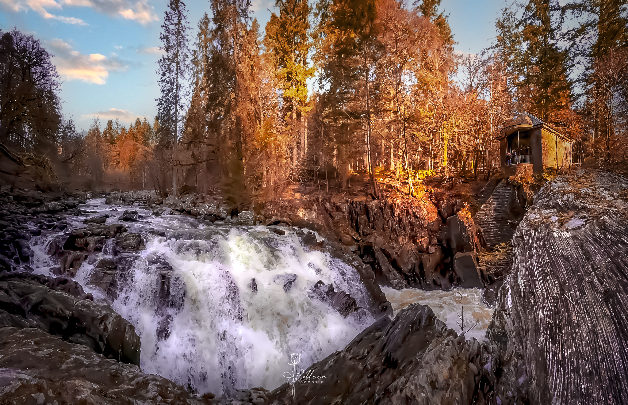 A scenic view of The Hermitage in Dunkeld, featuring a waterfall surrounded by trees and a structure in the distance.